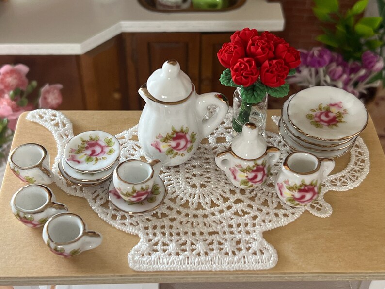 Może przedstawiać: Miniaturowy porcelanowy zestaw do herbaty z białym czajnikiem, dzbankiem na śmietanę, cukiernicą i czterema filiżankami i spodkami. Zestaw zdobiony jest kwiatowym wzorem z r&oacute;żowymi r&oacute;żami i zielonymi liśćmi. Zestaw do herbaty jest prezentowany na białym koronkowej serwetce z bukietem czerwonych r&oacute;ż w szklanym wazonie.