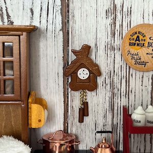 Op de afbeelding: Een miniatuur houten koekoeksklok met een vogelvormige bovenkant en slinger. De klok heeft een witte wijzerplaat met zwarte wijzers en cijfers. De scène omvat een miniatuurkeuken met een kast, koperen potten en vintage zuivelborden.