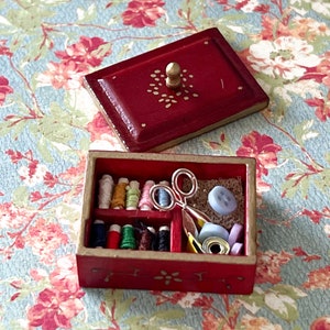 Puede incluir: Una caja de costura de madera roja con ribete dorado y un dise&ntilde;o floral. La caja est&aacute; abierta y contiene varios art&iacute;culos de costura, incluidos carretes de hilo, tijeras, botones y un enhebrador de agujas.