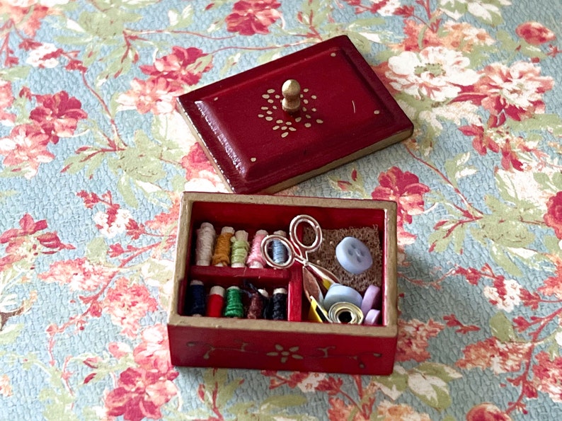 Puede incluir: Una caja de costura de madera roja con ribete dorado y un dise&ntilde;o floral. La caja est&aacute; abierta y contiene varios art&iacute;culos de costura, incluidos carretes de hilo, tijeras, botones y un enhebrador de agujas.
