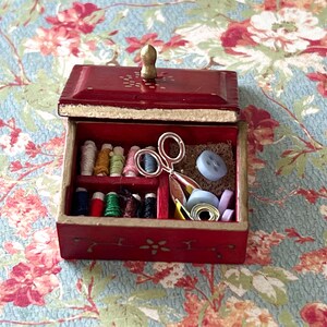 Puede incluir: Una peque&ntilde;a caja de madera roja con una tapa con bisagras y un pomo dorado. La caja est&aacute; abierta y contiene varios art&iacute;culos de costura, como carretes de hilo, botones y un par de tijeras.