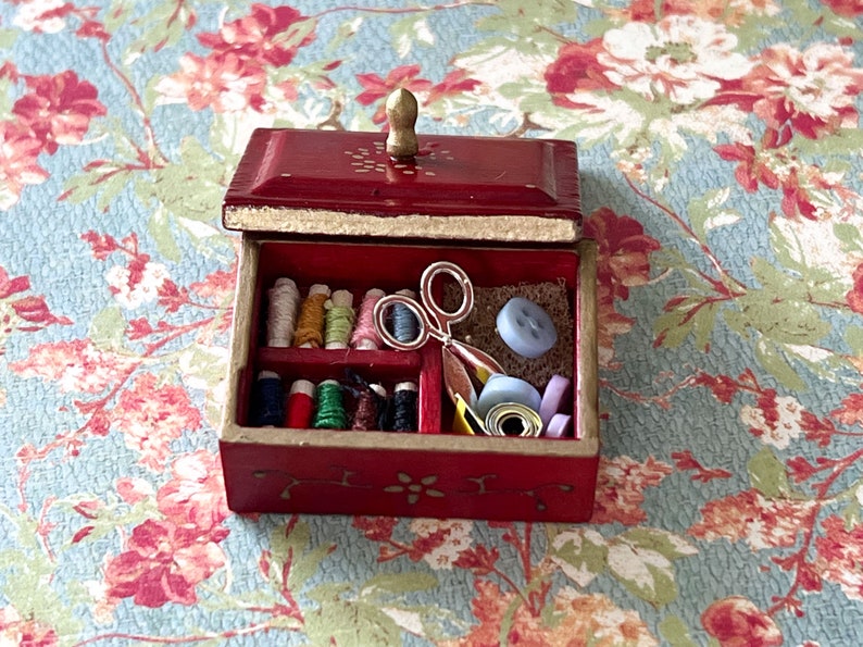 Puede incluir: Una peque&ntilde;a caja de madera roja con una tapa con bisagras y un pomo dorado. La caja est&aacute; abierta y contiene varios art&iacute;culos de costura, como carretes de hilo, botones y un par de tijeras.