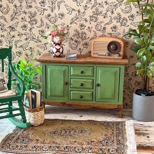 May include: A miniature room scene with a green rocking chair, a green cabinet, and a vintage radio. A small rug and a potted plant add to the cosy atmosphere. The wallpaper has a floral and bird pattern. A vase of flowers sits on the cabinet.