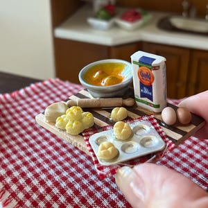 Pode incluir: Cena de cozinha em miniatura com uma toalha de mesa xadrez vermelha e branca. Inclui uma tábua de cortar com pãezinhos em miniatura, uma tigela de sopa, um saco de farinha Gold para todos os fins, um rolo e dois ovos. Uma assadeira em miniatura também está presente.