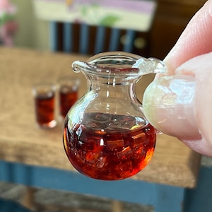 May include: A miniature clear glass pitcher filled with a red liquid and ice cubes. The pitcher has a small handle and a flared spout. Two small glasses with red liquid are in the background.