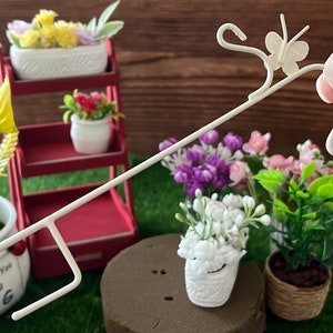 May include: White metal plant stand with a butterfly design. The stand is holding a small white pot with white flowers. The stand is on a brown surface with green grass.