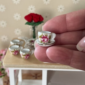 Może przedstawiać: Miniaturowy zestaw filiżanki i spodeczka do herbaty z kwiatowym wzorem. Filiżanka i spodeczek są białe z r&oacute;żowymi i zielonymi kwiatowymi akcentami. Spodeczek ma złotą obw&oacute;dkę.