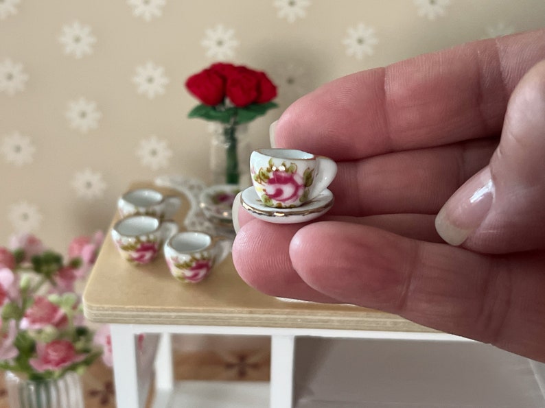 Może przedstawiać: Miniaturowy zestaw filiżanki i spodeczka do herbaty z kwiatowym wzorem. Filiżanka i spodeczek są białe z r&oacute;żowymi i zielonymi kwiatowymi akcentami. Spodeczek ma złotą obw&oacute;dkę.