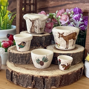 May include: Five miniature ceramic flower pots with different animal designs. The pots are arranged on wooden slices, creating a tiered display. The designs include a rabbit, a deer, an owl, a pine cone, and a squirrel.