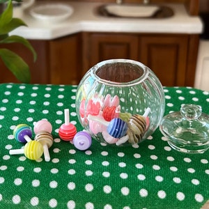 May include: A clear glass candy jar with a lid, filled with colourful candy. The jar is sitting on a green tablecloth with white polka dots. There are also several colourful lollipops on the tablecloth.