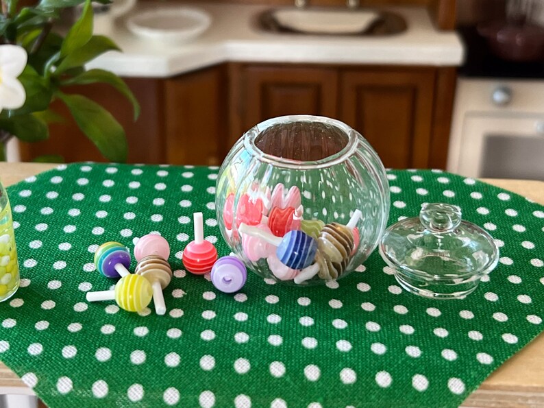 May include: A clear glass candy jar with a lid, filled with colourful candy. The jar is sitting on a green tablecloth with white polka dots. There are also several colourful lollipops on the tablecloth.