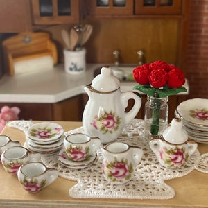 Może przedstawiać: Miniaturowy porcelanowy zestaw do herbaty z białą podstawą i wzorem r&oacute;żowych r&oacute;ż. Zestaw zawiera czajnik, dzbanek do śmietanki, cukiernicę, cztery filiżanki do herbaty i cztery spodki. Zestaw jest prezentowany na białym koronkowej serwetce z bukietem czerwonych r&oacute;ż w szklanym wazonie.