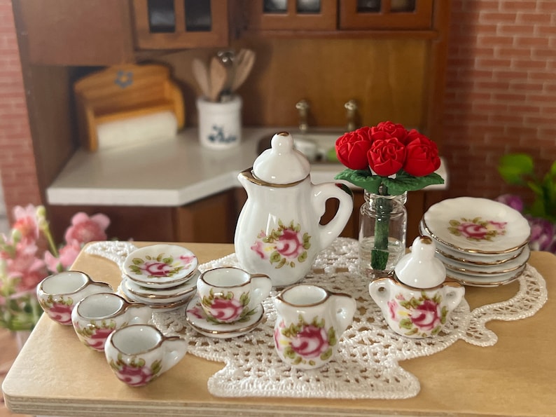 Może przedstawiać: Miniaturowy porcelanowy zestaw do herbaty z białą podstawą i wzorem r&oacute;żowych r&oacute;ż. Zestaw zawiera czajnik, dzbanek do śmietanki, cukiernicę, cztery filiżanki do herbaty i cztery spodki. Zestaw jest prezentowany na białym koronkowej serwetce z bukietem czerwonych r&oacute;ż w szklanym wazonie.