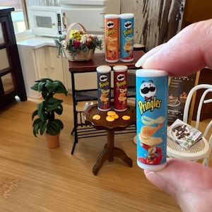 May include: Miniature Pringles potato chip cans in various flavors, including original, sour cream and onion, and paprika. The blue can is held by a person. Other miniature items include a table, plant, and a small cabinet.