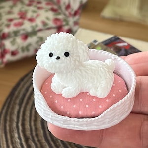 May include: A miniature white Bichon Frise dog figurine resting on a pink polka-dot cushion inside a white woven basket. The dog has black eyes and a fluffy coat. This decorative item is likely for a dollhouse or miniature scene.