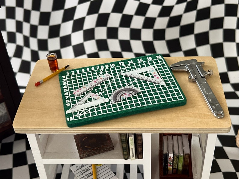 May include: A green cutting mat with a grid pattern and numbers 1-20, a protractor, two rulers, and a silver caliper on a wooden table with a shelf.