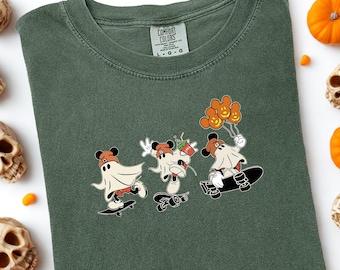 May include: Moss green t-shirt featuring a Halloween-themed graphic of three cartoon characters on skateboards, dressed as ghosts. The shirt is surrounded by decorative skulls, pumpkins, and candy corn. The words "COMFORT COLORS" and "MOSS" are also visible.