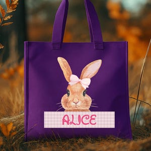 May include: A purple tote bag featuring a watercolor illustration of a bunny with a pink bow. The bunny is centered on the bag, above a pink and white checkered banner with the name "ALICE" in pink script.
