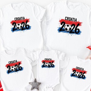 May include: White t-shirts with a Croatia soccer design. The design includes the word "Croatia", the number "2026", and a soccer ball, with red, white, and blue accents. The shirts are available in various sizes.