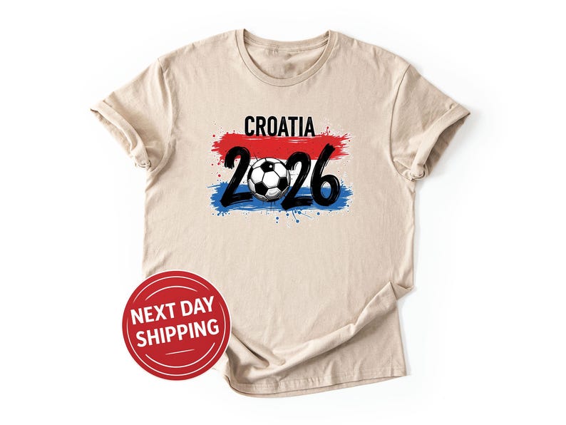 May include: Beige t-shirt with the word "CROATIA" in black above a red and blue paint stroke design. Below the design is the number "26" in black with a soccer ball in the middle. A red circle with the text "NEXT DAY SHIPPING" is in the bottom left.