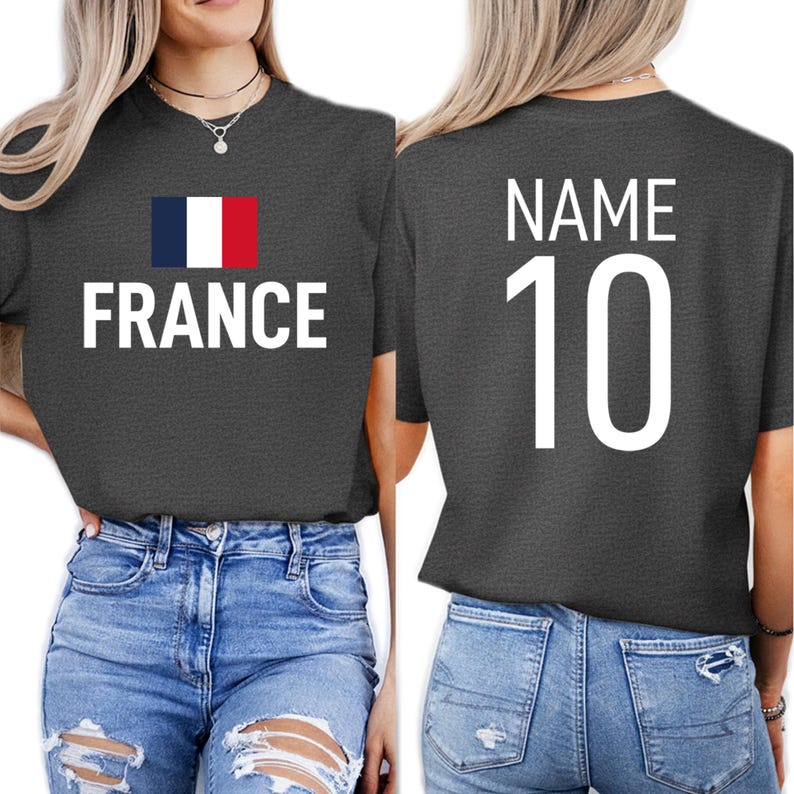 May include: Dark grey t-shirt featuring the word "FRANCE" in white block letters and the French flag. The back of the shirt displays "NAME" above the number "10" in white. The shirt is worn with blue jeans.