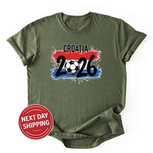 May include: Olive green t-shirt with a graphic featuring the word "CROATIA" above the numbers "26" and a soccer ball. The design incorporates the colors of the Croatian flag: red, white, and blue. A red circle with the text "NEXT DAY SHIPPING" is at the bottom.