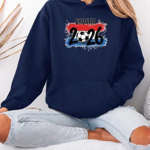 May include: Navy blue hooded sweatshirt featuring the word "CROATIA" above the number "2026" and a soccer ball graphic. The design incorporates the colors of the Croatian flag: red, white, and blue. The sweatshirt has a front pocket and a drawstring hood.