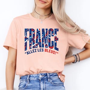 May include: A peach-colored t-shirt with the word "FRANCE" in large, blue and red letters, filled with French-themed illustrations. Below the word "FRANCE" is the phrase "ALLEZ LES BLEUS!" in white text.