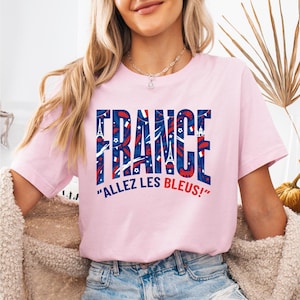 May include: A light pink t-shirt with the word "FRANCE" in large, patriotic lettering. The letters are filled with red, white, and blue patterns, including the Eiffel Tower and stars. Below the word "FRANCE" is the phrase "ALLEZ LES BLEUS!"
