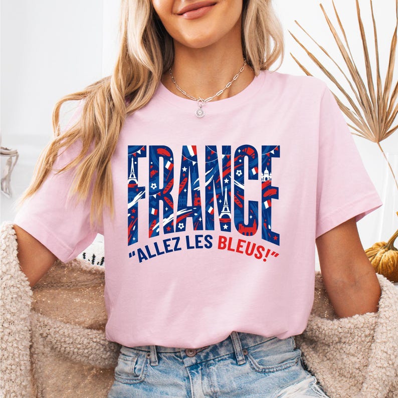 May include: A light pink t-shirt with the word "FRANCE" in large, patriotic lettering. The letters are filled with red, white, and blue patterns, including the Eiffel Tower and stars. Below the word "FRANCE" is the phrase "ALLEZ LES BLEUS!"