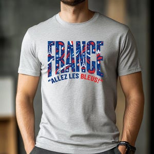 May include: A light gray t-shirt with the word "FRANCE" in blue and red, filled with French-themed graphics. Below the word "FRANCE" is the phrase "ALLEZ LES BLEUS!" in white. The shirt is short-sleeved.
