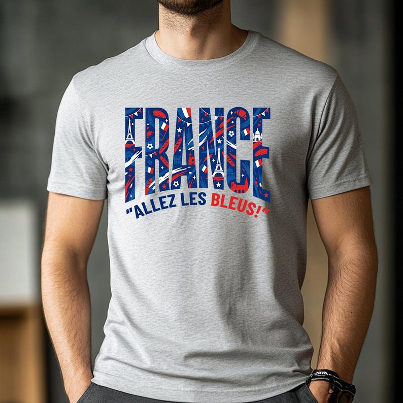 May include: A light gray t-shirt with the word "FRANCE" in blue and red, filled with French-themed graphics. Below the word "FRANCE" is the phrase "ALLEZ LES BLEUS!" in white. The shirt is short-sleeved.