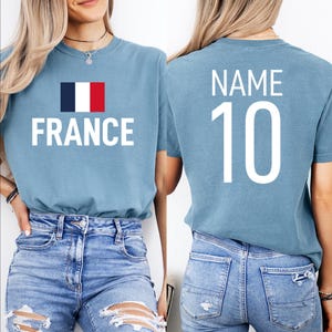 May include: Pale blue t-shirt featuring the word "FRANCE" in white, with the French flag. The back of the shirt displays "NAME" above the number "10" in white. The shirt is made of a soft material.