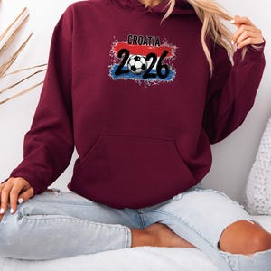 May include: A maroon hoodie with a front pocket and a drawstring hood. The front features the text "CROATIA 2026" with a soccer ball graphic, set against a red, white, and blue background, representing the Croatian flag.