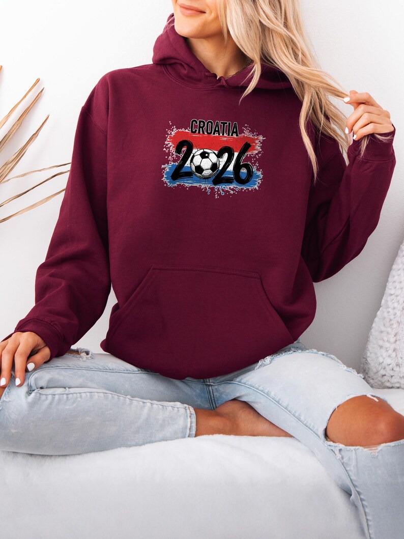 May include: A maroon hoodie with a front pocket and a drawstring hood. The front features the text "CROATIA 2026" with a soccer ball graphic, set against a red, white, and blue background, representing the Croatian flag.