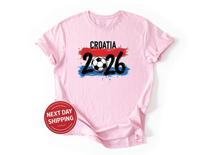 May include: A light pink t-shirt with the word "CROATIA" above a soccer ball and the numbers "2026". The design incorporates red, white, and blue brushstrokes, and a soccer ball. The shirt has a round neck and short sleeves.