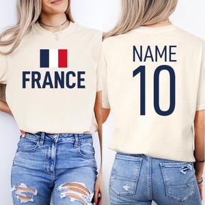 May include: Cream-coloured t-shirt featuring the word "FRANCE" in navy blue, accompanied by the French flag. The back of the shirt shows "NAME" above the number "10" in navy blue. The shirt is worn with blue jeans.