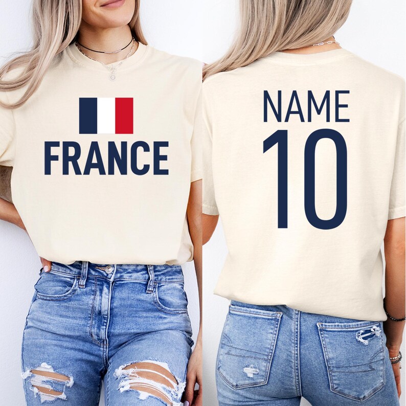 May include: Cream-coloured t-shirt featuring the word "FRANCE" in navy blue, accompanied by the French flag. The back of the shirt shows "NAME" above the number "10" in navy blue. The shirt is worn with blue jeans.