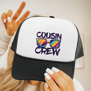May include: A black and white trucker hat with the words "COUSIN CREW" and a graphic of sunglasses in vibrant colors. The hat has a white front panel and a black mesh back.