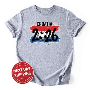 May include: A gray t-shirt with the word "CROATIA" above the number "2026" and a soccer ball graphic. The design incorporates red, white, and blue brushstrokes, resembling a flag. The shirt is made of a soft material.