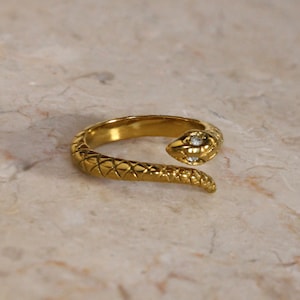 May include: A gold snake ring with a single clear stone on the head. The ring is textured and has a detailed design.