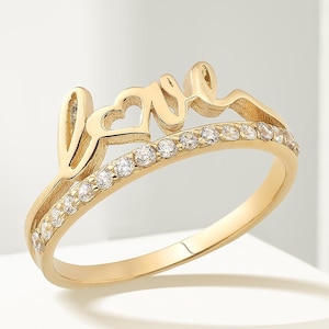 Op de afbeelding: Een goudkleurige ring met het woord "love" in cursief schrift. De ring heeft een band van kleine, heldere stenen en een hartvormig detail in het "love"-ontwerp. De ring wordt op een wit oppervlak gepresenteerd.
