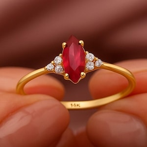 May include: A gold ring featuring a marquise-cut ruby gemstone, flanked by small, round diamonds. The ring's band is slender and gold-colored. The ruby is a vibrant red color, and the diamonds sparkle. The ring is held between two hands.