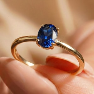 Könnte beinhalten: Ein goldener Ring mit einem leuchtend blauen, ovalen Edelstein. Der Ring hat ein poliertes Goldband, und der Edelstein ist das Hauptmerkmal, das von vier Krappen gehalten wird. Der Ring wird von einer Person gehalten.