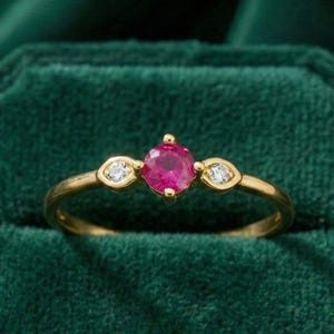 May include: A gold ring with a central round ruby gemstone and two smaller, oval-shaped diamonds on either side. The ring is set against a dark green velvet background, highlighting the vibrant colors of the gemstones.
