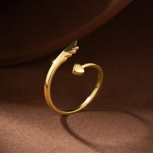May include: A gold-colored ring with a wing design and a heart-shaped accent. The ring has a delicate, open design, with the wing extending from one side and the heart on the other. The ring is set against a brown background.