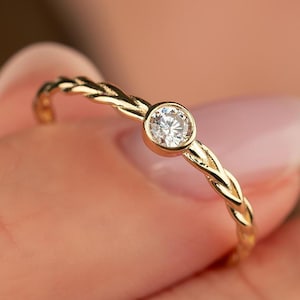 May include: A gold ring featuring a braided band and a single, round, clear gemstone. The ring is held by fingers, highlighting the delicate design and the sparkle of the stone. The ring is a piece of jewellery.