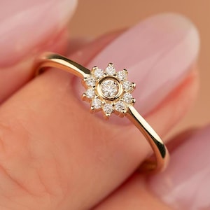 Könnte beinhalten: Ein zarter goldener Ring mit floralem Design, mit einem zentralen Diamanten, der von kleineren Steinen umgeben ist. Der Ring hat ein dünnes, elegantes Band, das einen minimalistischen Stil zeigt. Der Ring wird am Finger einer Person gezeigt.