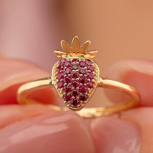 May include: A gold-colored ring featuring a strawberry design. The strawberry is made of small, round, red gemstones. The ring has a delicate band and a crown-like detail at the top of the strawberry. The ring is held between two fingers.