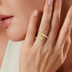 May include: A gold-colored ring with an open design. One end features a wing shape, while the other has a heart-shaped point. The ring is worn on a finger, showcasing its delicate details and elegant style.